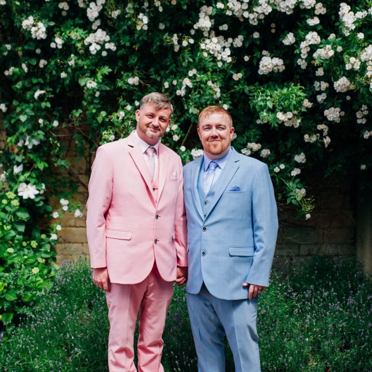 Two men in pastel suits stand smiling in front of flowering greenery.